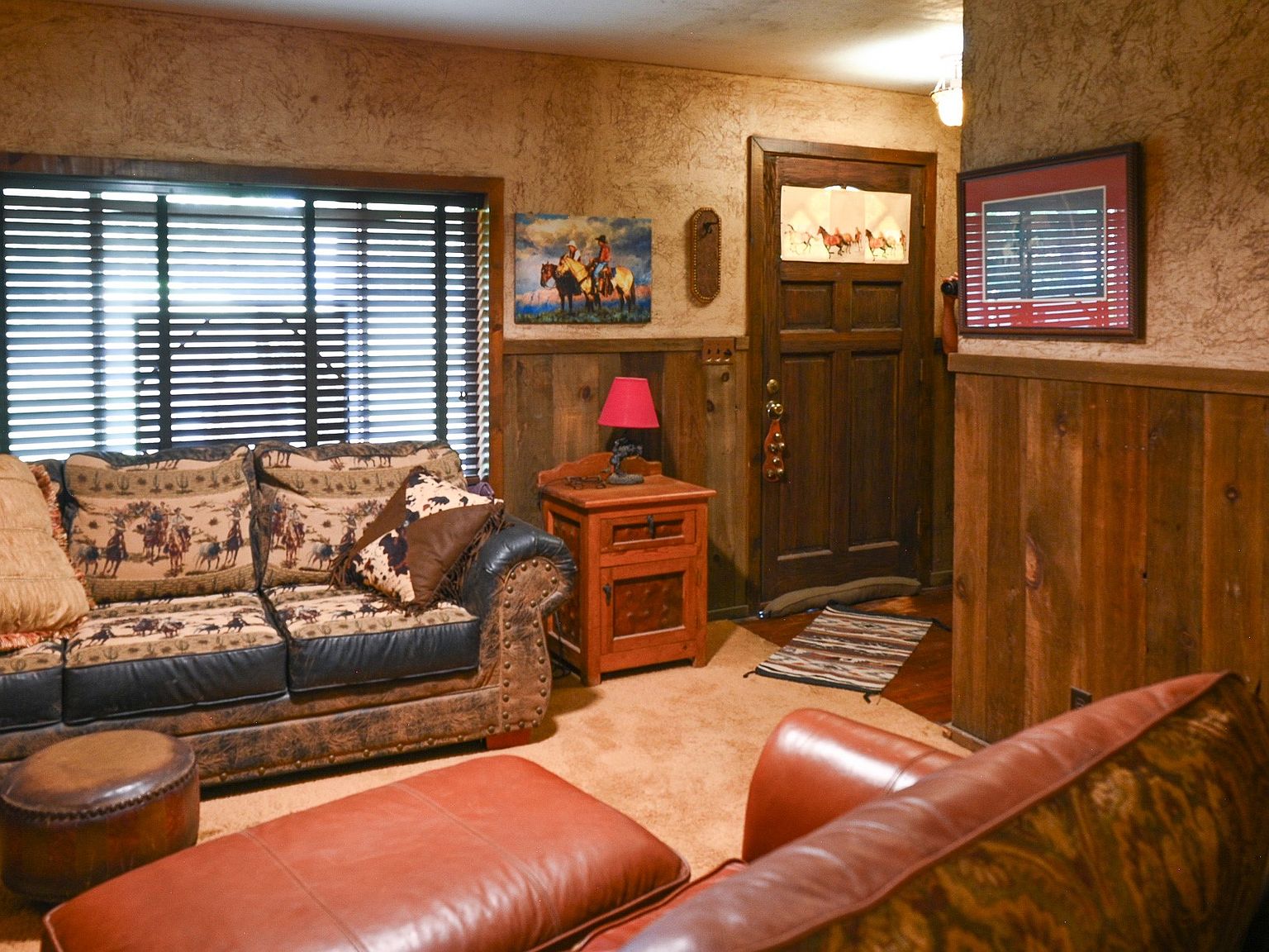 Western-themed living room with leather and cowboy-print sofas, knotty pine wainscoting, framed equestrian artwork, and bright daylight through plantation blinds