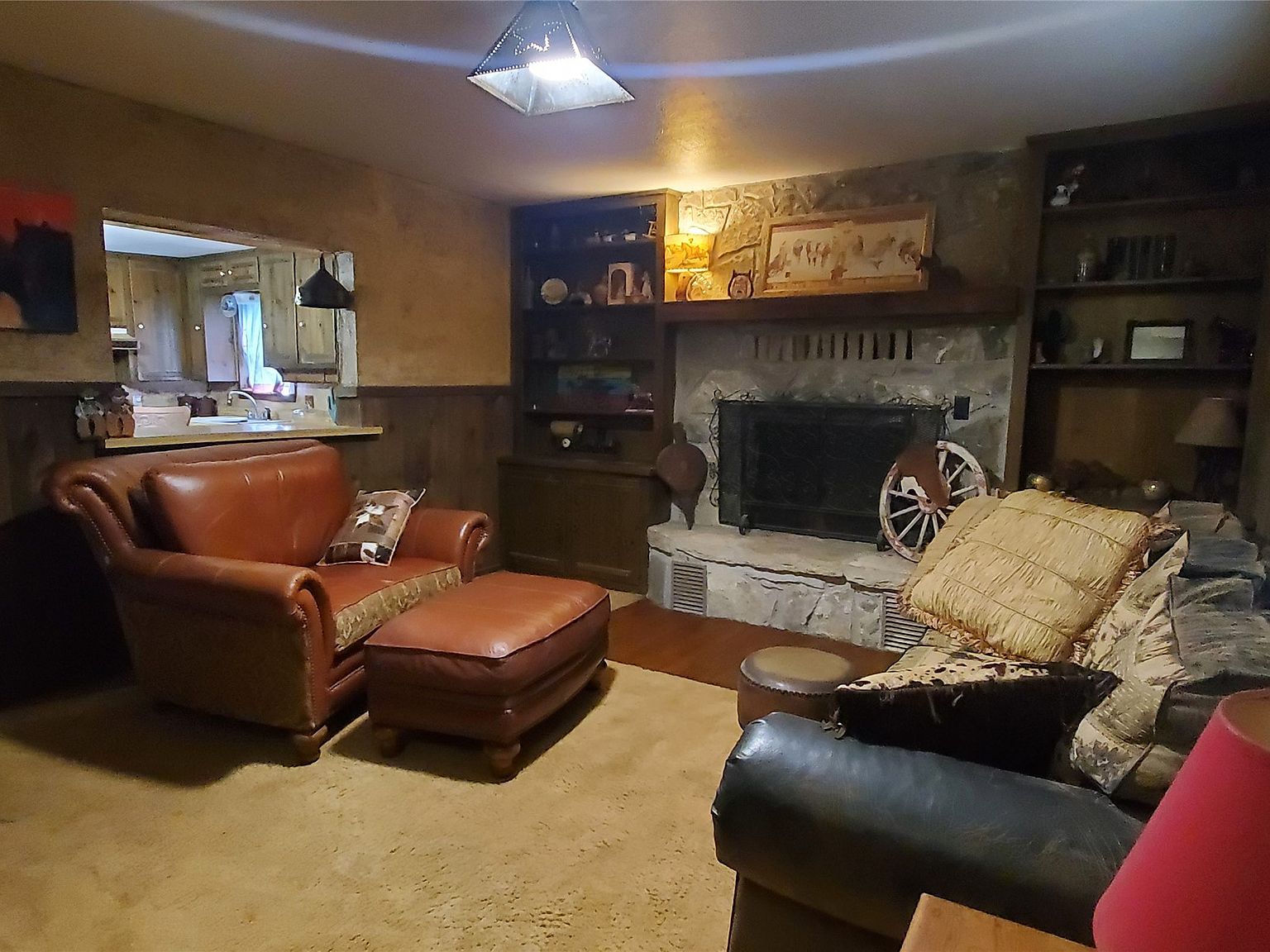 Same great room from the kitchen pass-through angle: stone fireplace at the right, leather club chair and ottoman at the center, kitchen counter visible at the left