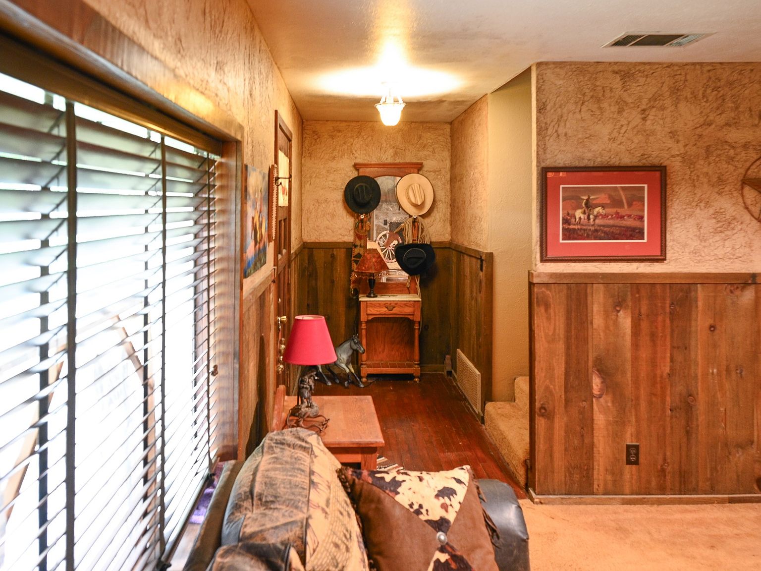 Narrow entry hallway with knotty pine wainscoting, a wooden console table topped by cowboy hats hung on the wall, and warm overhead lamp light