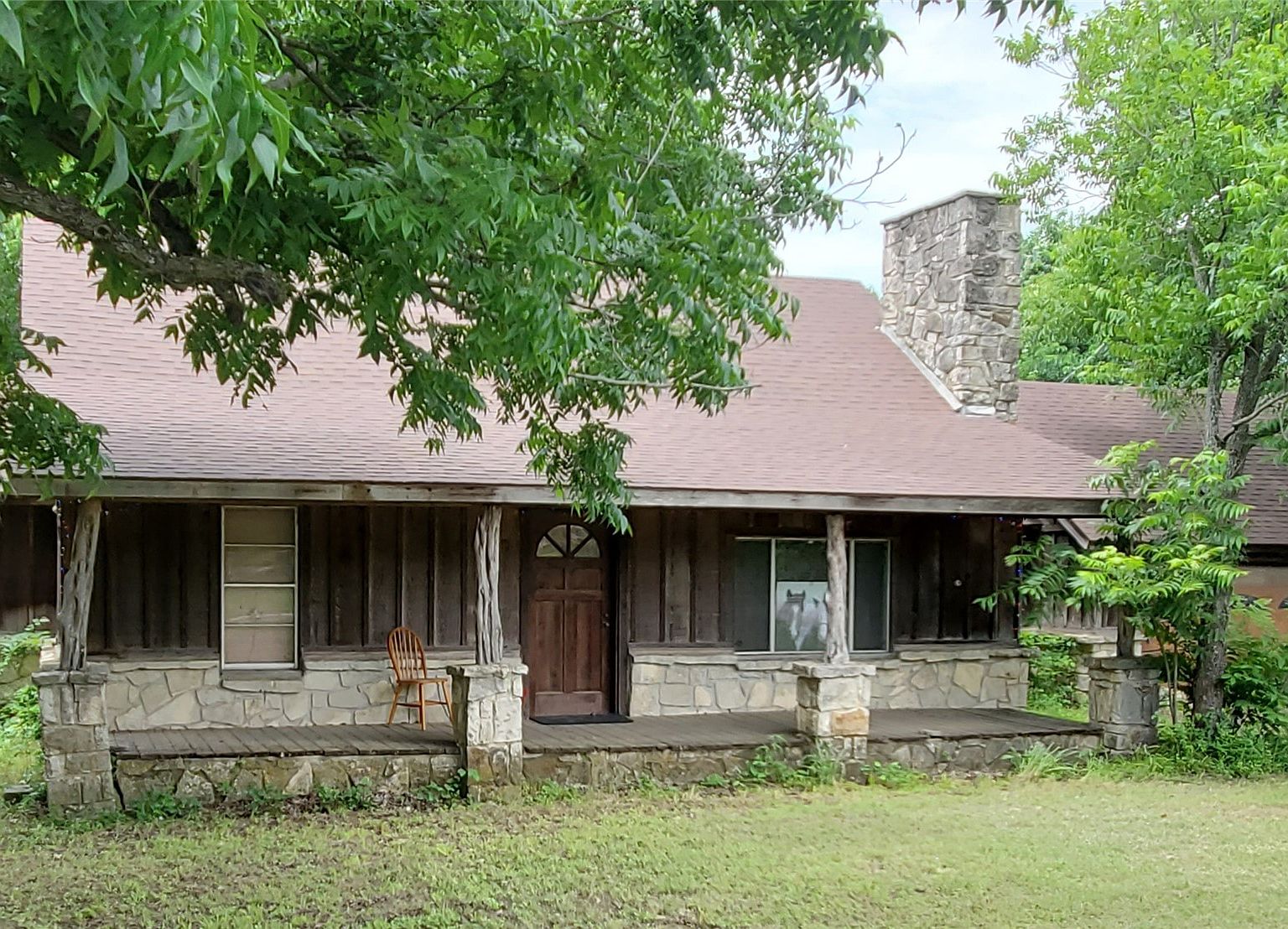 Front elevation of the main house at 8401 FM 2738: a single-story stone-and-cedar country home with a covered porch, framed by mature pecan and oak trees